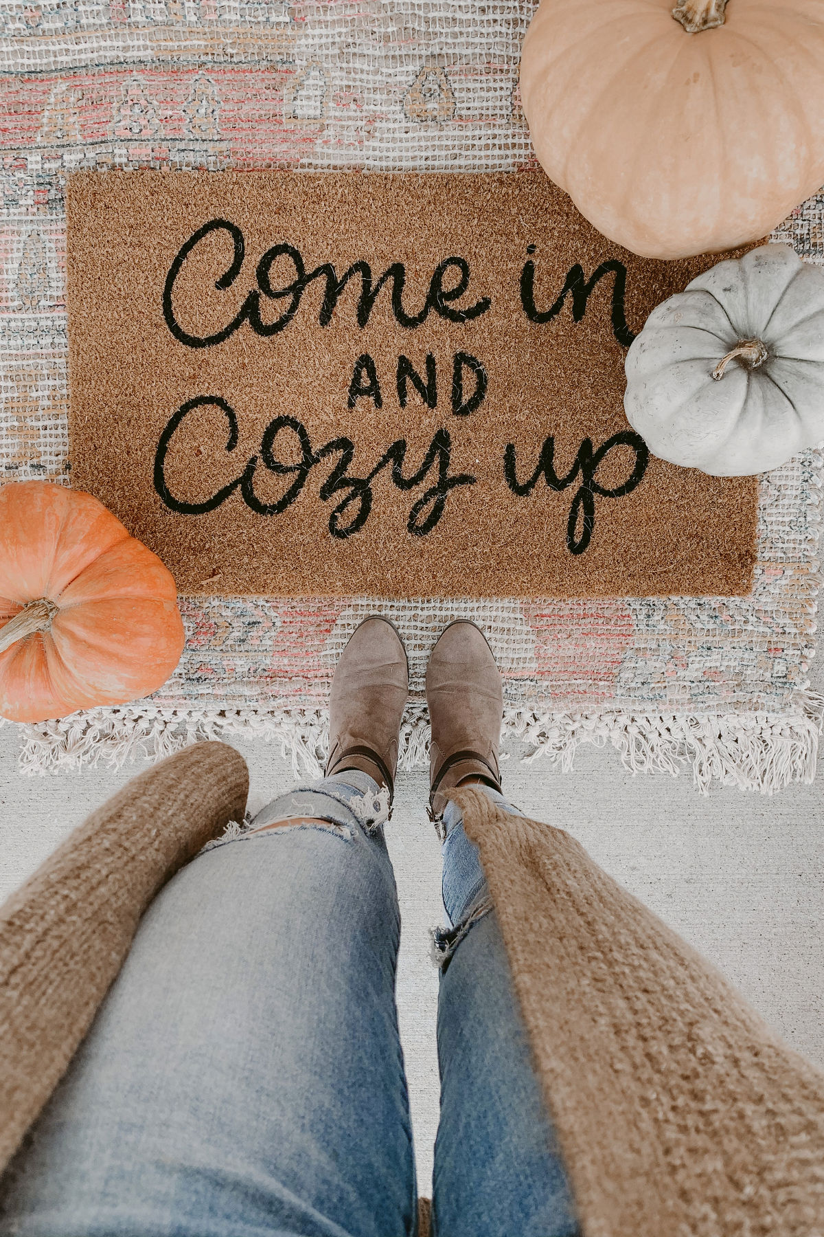 POV of a woman looking down at her feet in front of a door mat surrounded by pumpkins that say 'come in and cozy up'